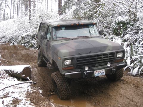 1989 Ford Bronco - A camouflage Ford Bronco is shown from a 3/4 front angle, navigating a muddy, snow-dusted trail through a forest. The vehicle is equipped w…