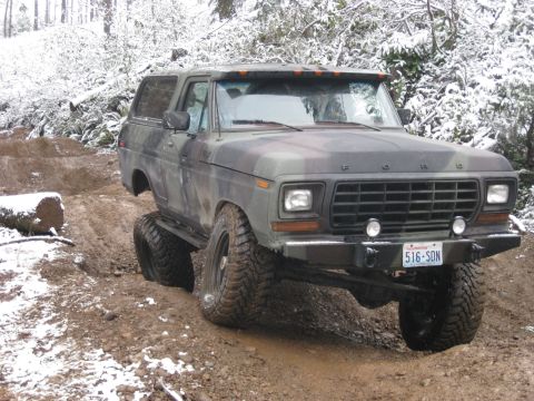 1989 Ford Bronco - A camouflage-painted Ford Bronco is navigating a muddy, snow-dusted off-road trail. The vehicle is angled to the left, with its front right…
