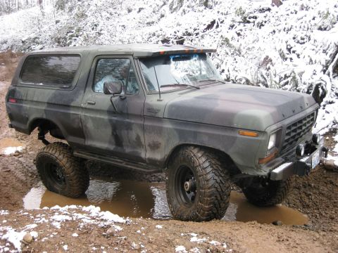 1989 Ford Bronco - A camouflage-painted Ford Bronco is navigating a muddy off-road trail with snow on the surrounding trees and ground.