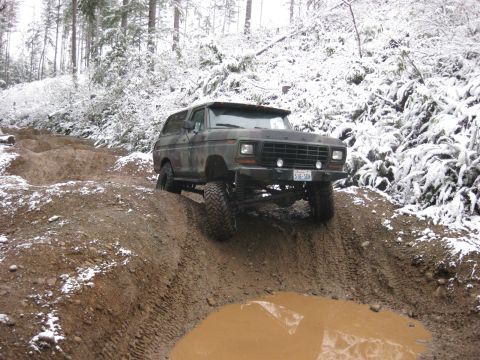 1989 Ford Bronco - A lifted, dark green Ford Bronco is navigating a muddy, snow-dusted off-road trail. The vehicle is angled upwards on a dirt embankment, wit…