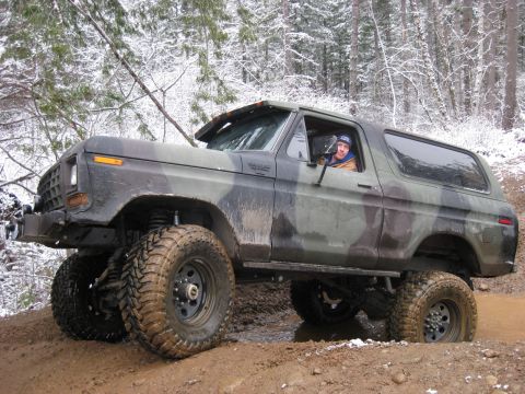 1989 Ford Bronco - A camouflage-painted Ford Bronco is shown navigating a muddy, snow-dusted trail in a forest. The driver is visible behind the wheel, lookin…