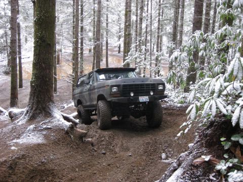 1989 Ford Bronco - A dark-colored Ford Bronco is navigating a muddy, wooded trail with light snow on the ground and trees. The vehicle is angled towards the v…