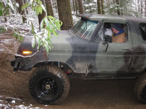 1989 Ford Bronco - A camouflage-painted Ford Bronco drives off-road through a snowy forest. The vehicle is muddy, and branches with snow are visible in the fo…