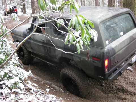 1989 Ford Bronco - A dark green Ford Bronco is driving off-road through a snowy, wooded area. The vehicle is tilted on a muddy incline, with snow-covered bran…