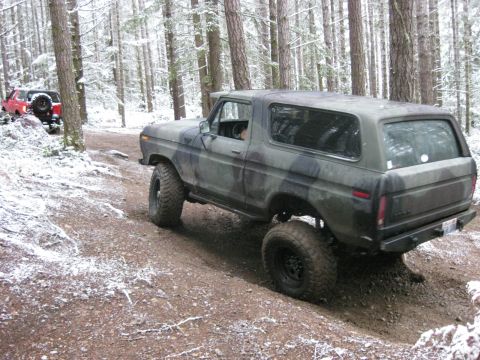 1989 Ford Bronco - A camouflage-painted Ford Bronco is driving on a snowy, wooded trail. Another red SUV is visible in the background.