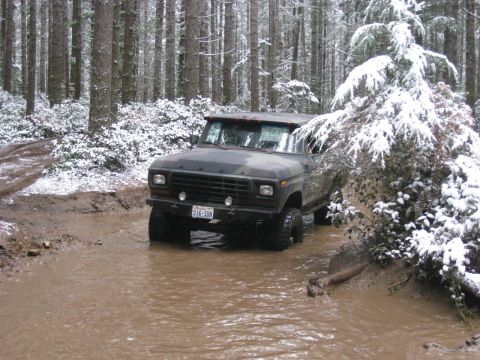1989 Ford Bronco - A dark green camouflage Ford Bronco is driving through a muddy stream in a snowy forest. The vehicle is partially submerged in the water, w…