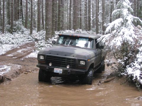 1989 Ford Bronco - A camouflaged Ford Bronco is driving through a muddy, snow-dusted forest road. The vehicle is partially submerged in the water, with snow v…