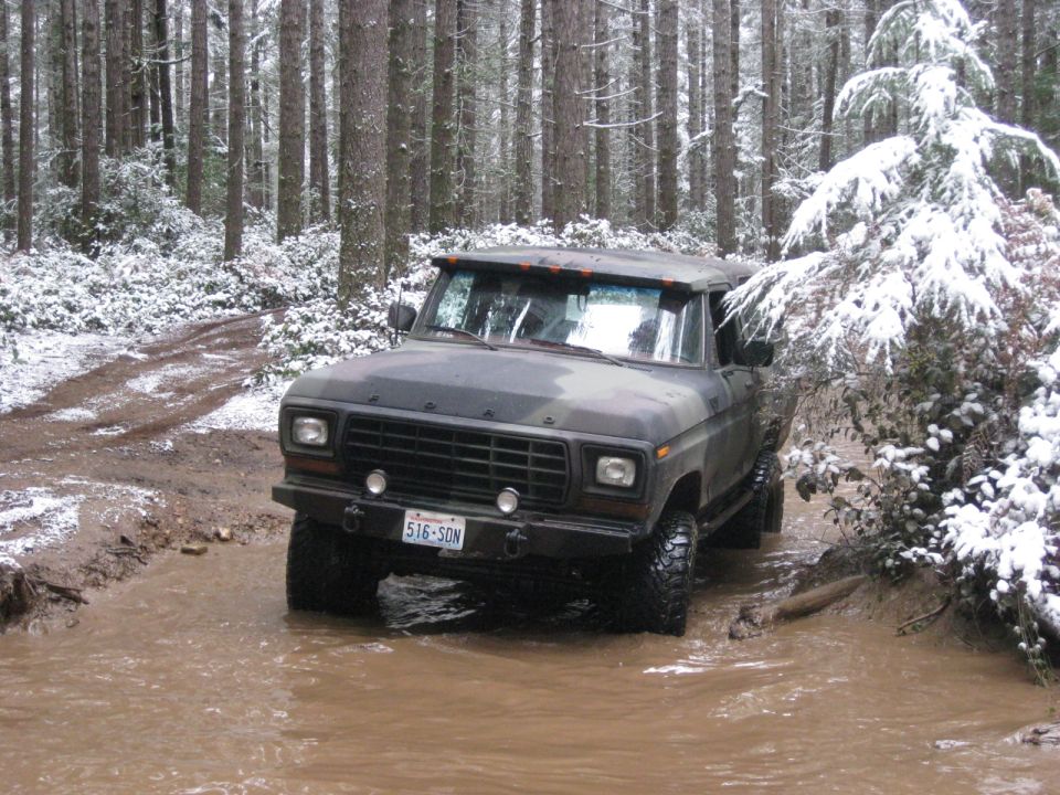 Camouflaged Ford Bronco driving through a muddy, snow-covered forest road.