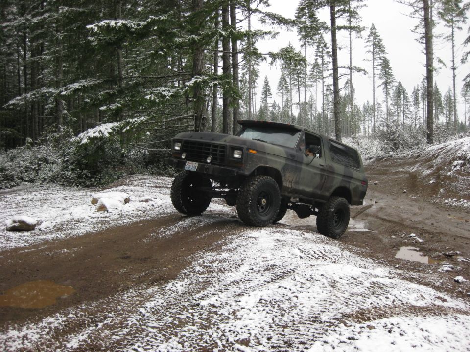 Lifted camouflage Ford Bronco driving on a snowy, muddy forest road.