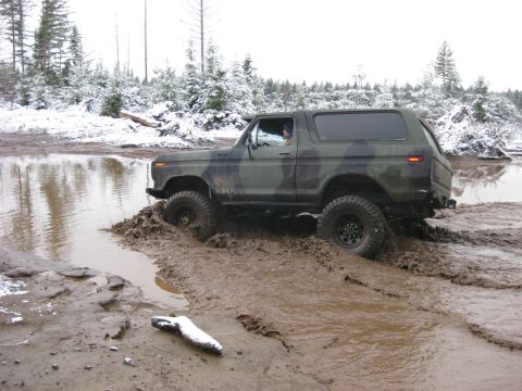 1989 Ford Bronco - A camouflage Ford Bronco is stuck in deep mud and water, with snow-covered trees in the background.