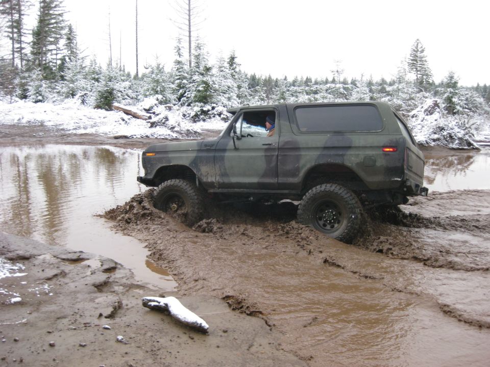 Camouflage Ford Bronco stuck in mud and water with snow on trees.