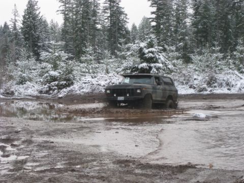 1989 Ford Bronco - A dark-colored Ford Bronco is driving through a muddy, flooded area with snow-covered trees in the background.