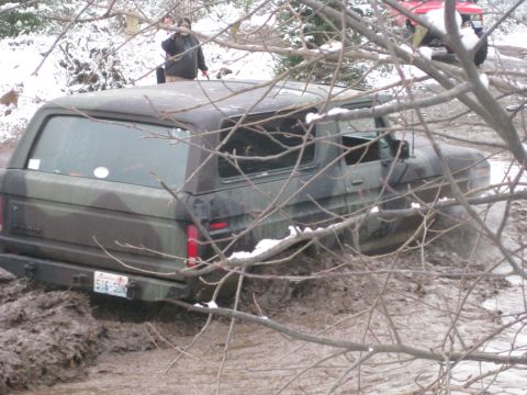 1989 Ford Bronco - A camouflage Ford Bronco is stuck in deep mud with snow on the ground and bare tree branches in the foreground. The rear of the vehicle sho…