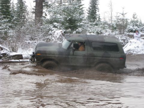 1989 Ford Bronco - A camouflage-painted Ford Bronco is driving through deep mud and water with snow-covered trees in the background. A person in a white jacke…