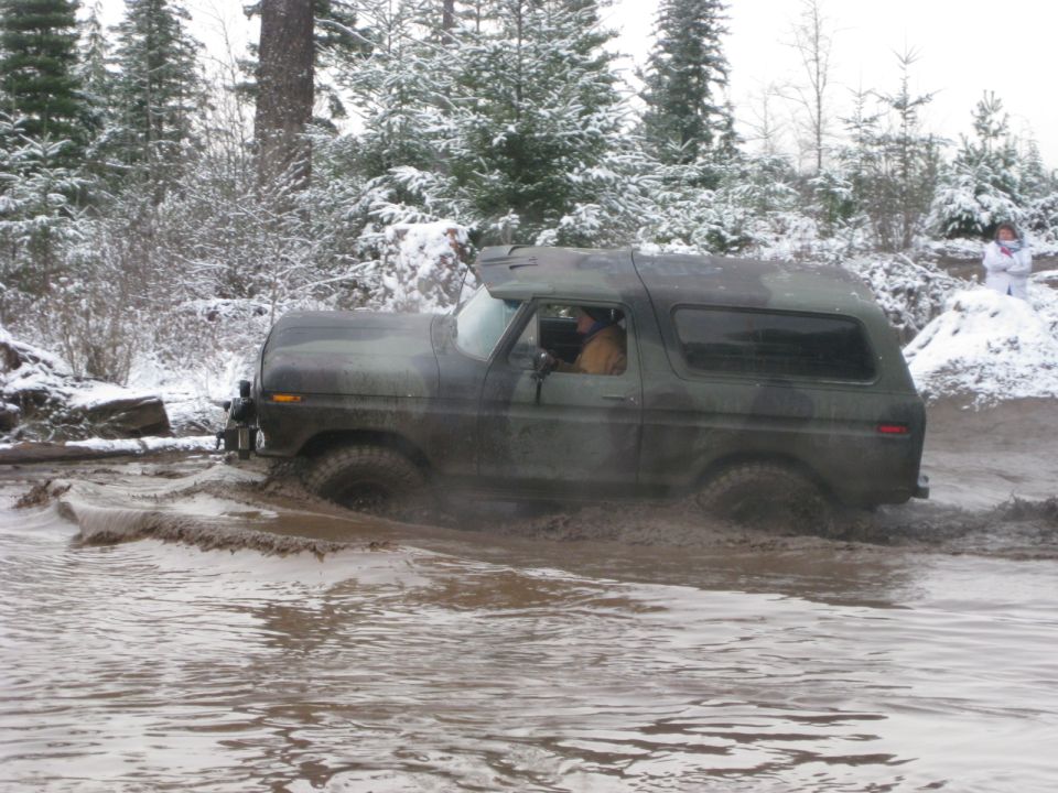Camouflage Ford Bronco driving through mud and water in a snowy forest.
