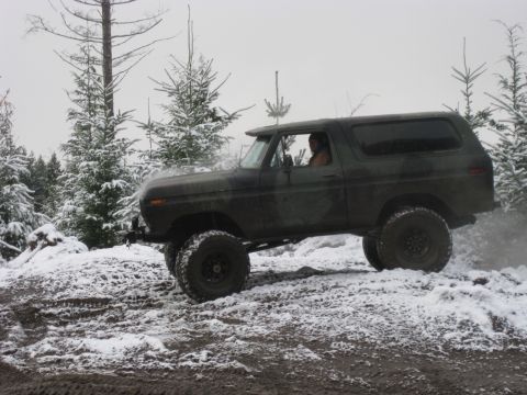 1989 Ford Bronco - A dark green, lifted Ford Bronco drives off-road through a snowy, wooded area. The vehicle is angled to the right, showing its side profile…