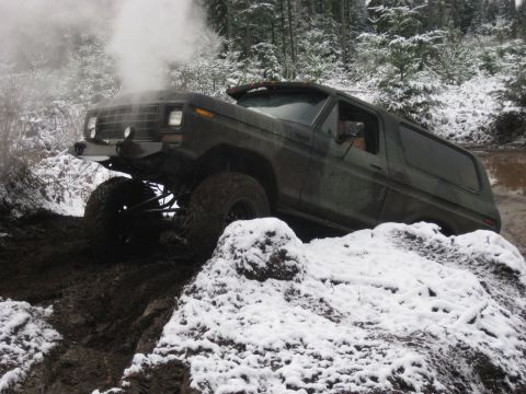 1989 Ford Bronco - A dark green Ford Bronco is navigating a muddy, snowy trail, with steam or smoke rising from the hood. The vehicle is angled upwards, with…