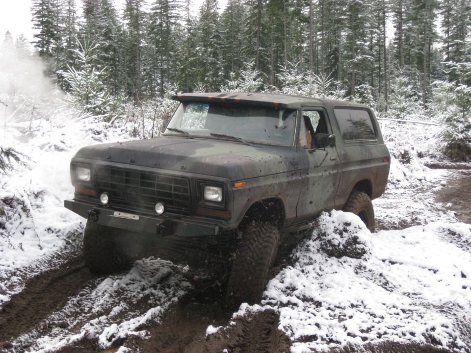 Camouflaged Ford Bronco driving in a snowy, muddy forest.