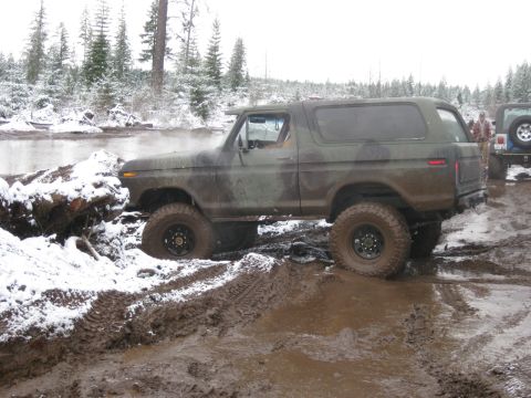 1989 Ford Bronco - A camouflage-painted Ford Bronco is navigating a muddy, snow-covered terrain next to a body of water. Another Jeep is visible in the backgr…