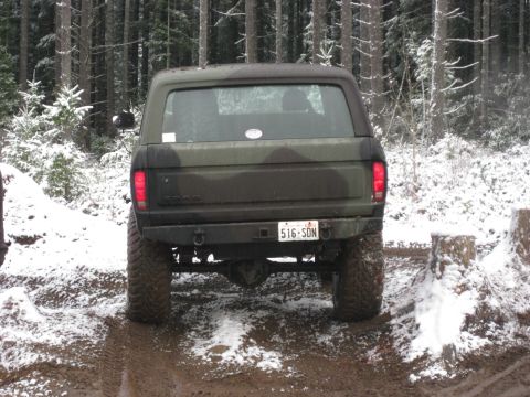 1989 Ford Bronco - The rear view of a camouflaged Ford Bronco parked on a muddy, snow-dusted trail in a forest. The vehicle has large off-road tires and a lic…