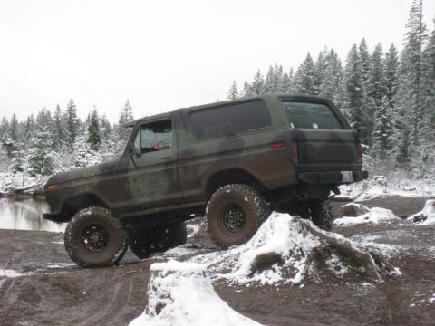 1989 Ford Bronco - A camouflage-painted Ford Bronco is shown in a muddy, snow-covered forest setting with a body of water to the left.