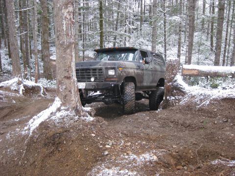 1989 Ford Bronco - A lifted, camouflage-painted Ford Bronco is navigating a muddy trail through a snowy forest. The vehicle is angled towards the viewer, show…