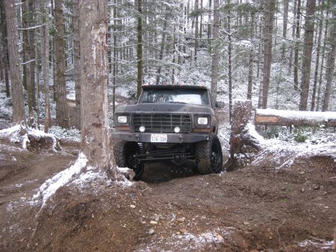 1989 Ford Bronco - A lifted, dark-colored Ford Bronco navigates a muddy, wooded trail with snow on the ground and trees. The front of the vehicle is visible,…