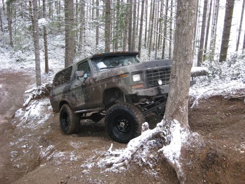 1989 Ford Bronco - A lifted, camouflage-painted Ford Bronco is shown navigating a muddy, snow-dusted forest trail. The vehicle is angled towards the viewer's…