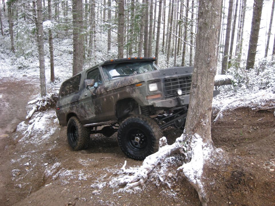 Lifted camouflage Ford Bronco on a muddy, snow-dusted forest trail, angled left, partially behind a tree.