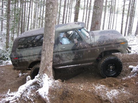 1989 Ford Bronco - A camouflaged Ford Bronco is navigating a muddy, wooded terrain with snow on the ground and trees surrounding it. The vehicle is angled on…
