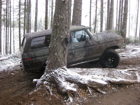 1989 Ford Bronco - A muddy, camouflage-painted 1980s Ford Bronco is parked on a dirt trail in a snowy forest, angled towards a large tree.