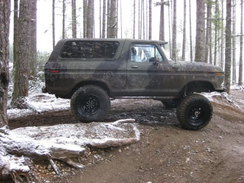1989 Ford Bronco - A muddy, lifted 1978-1979 Ford Bronco with aftermarket wheels and tires is parked on a dirt trail in a forest with snow on the ground.