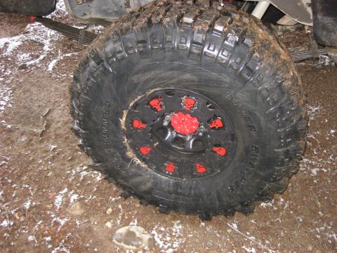 1989 Ford Bronco - A close-up of a large, off-road tire on a vehicle, featuring a black rim with red skull accents and a red center cap. The tire is mounted o…