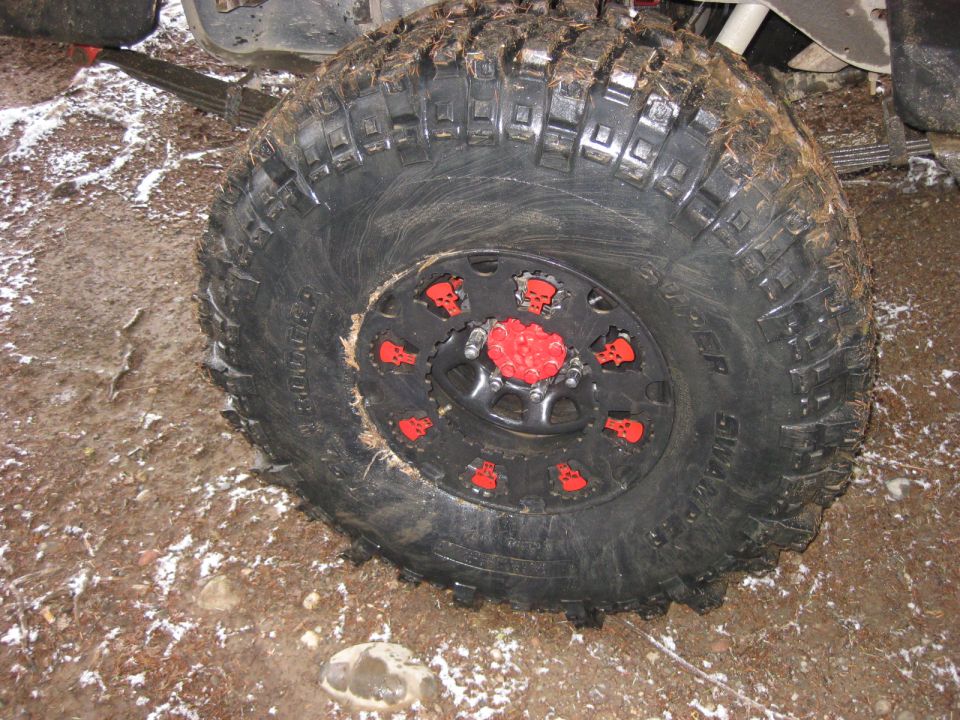 Off-road tire with black rim, red skull accents, and red center cap on a muddy surface.