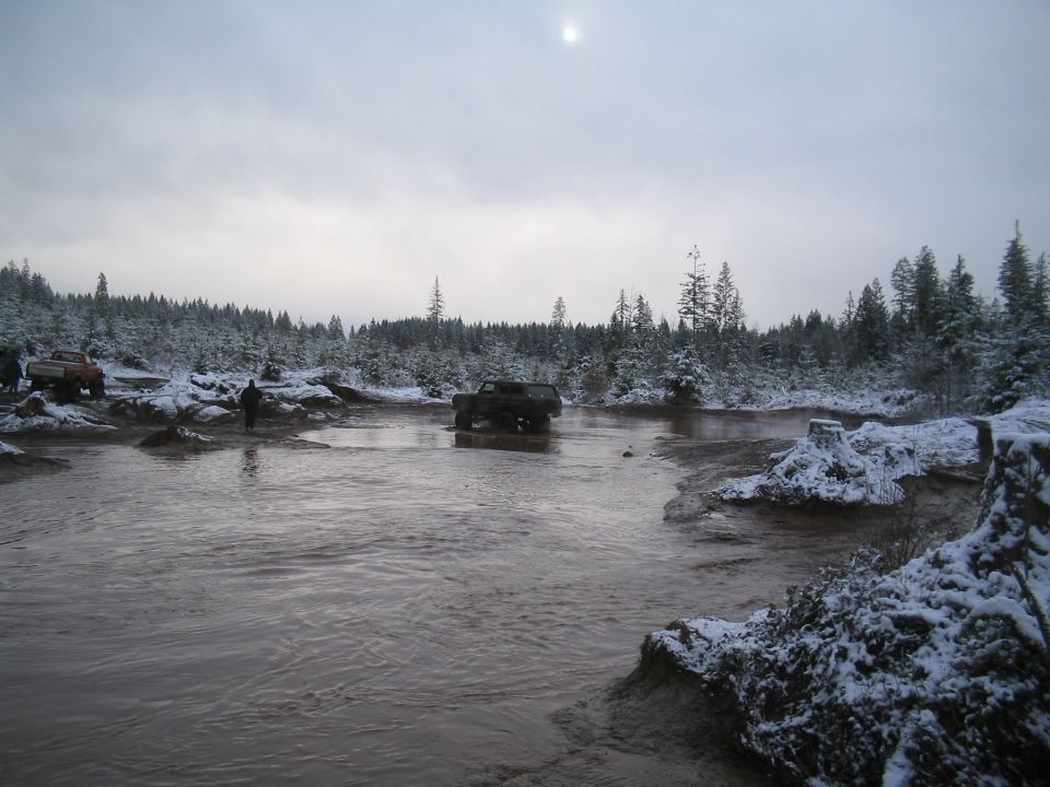 Dark SUV driving through muddy water with snow-covered trees and a red pickup in the background