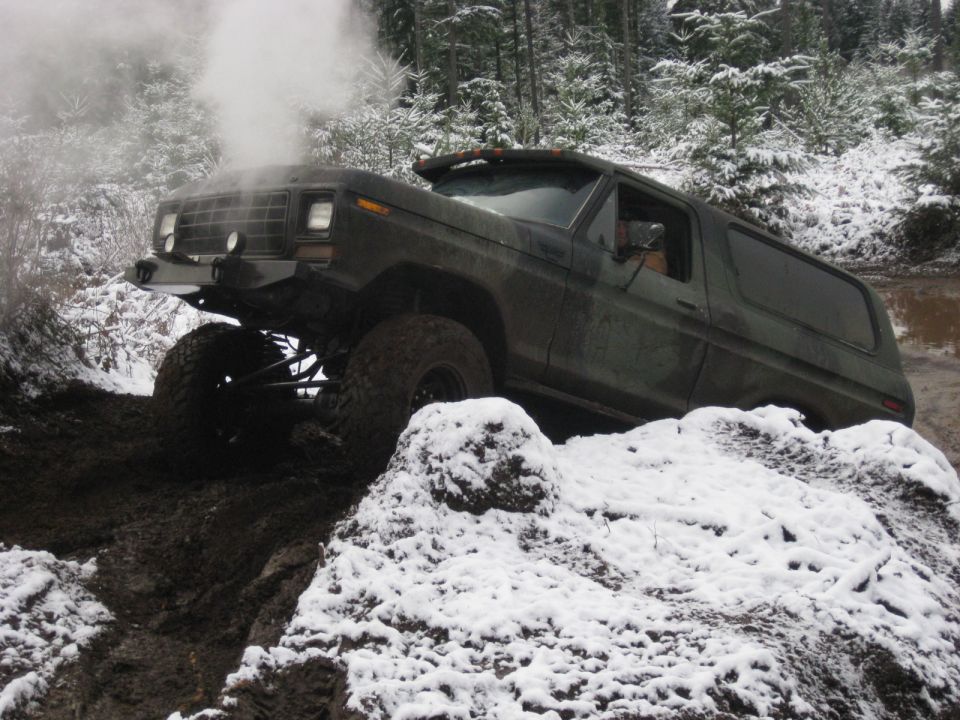 Dark green Ford Bronco off-roading on a muddy, snowy trail, steam rising from the engine.