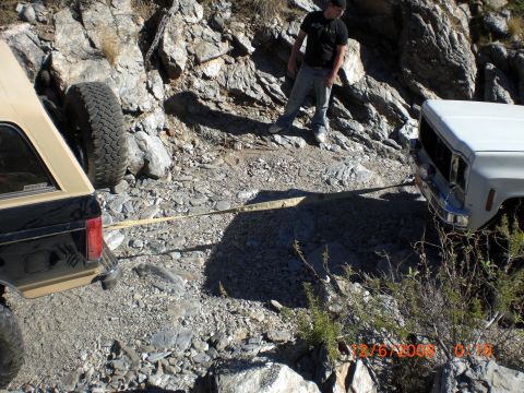 1991 Ford Bronco - A tan and black SUV and a white pickup truck are stuck on a rocky trail, connected by a tow strap.