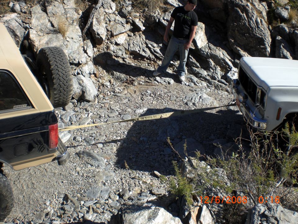 Two off-road vehicles, one tan and black SUV and one white pickup truck, are stuck on a rocky trail with a tow strap connecting them