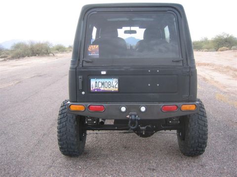 1987 Suzuki Samurai - Rear view of a black SUV with oversized tires and a custom bumper, parked on a dirt road.
