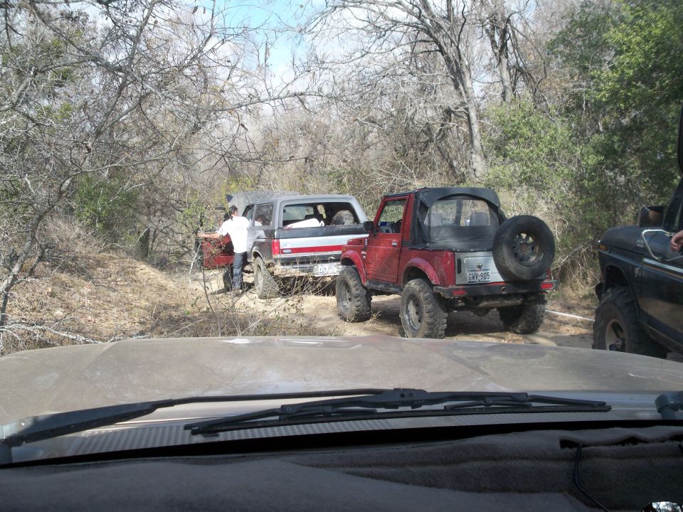View through windshield of trail with red-and-white lifted SUV and red soft-top 4x4 on mud tires