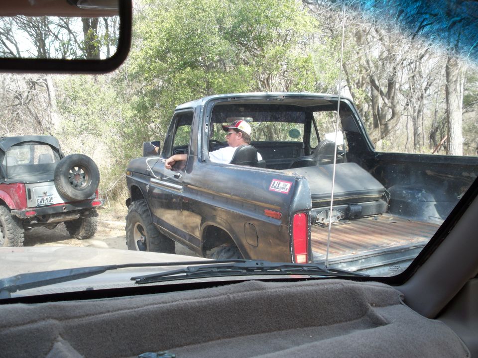 Topless black square-body 4x4 with bare bed floor and driver in cap on a wooded trail