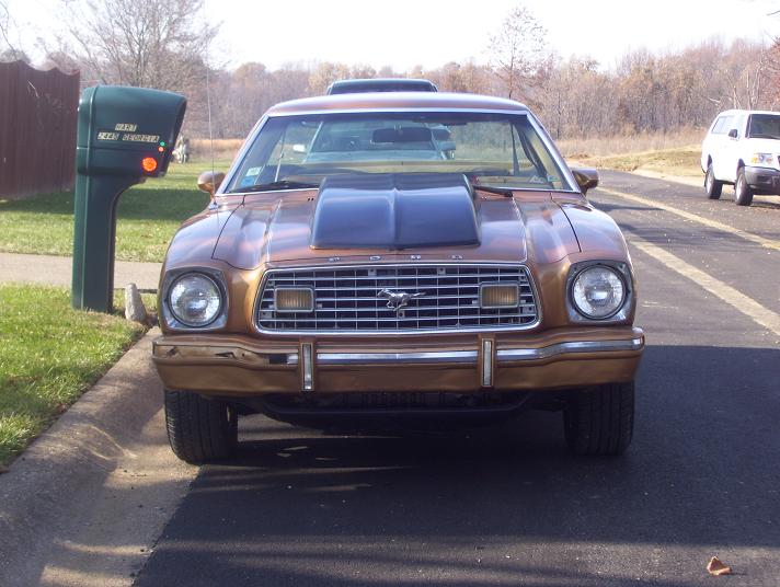 1977 Ford Mustang II with a hood scoop parked curbside, another SUV visible on the road