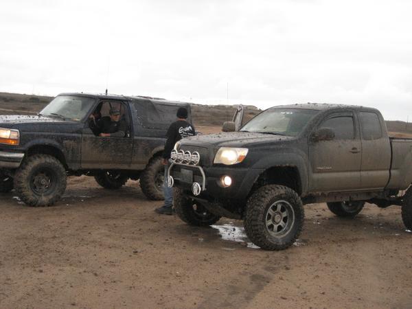 Two modified off-road trucks, a Ford Bronco and a Toyota Tacoma, parked on a dirt path.