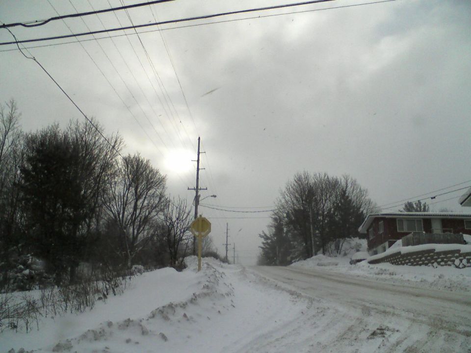 Snow-covered road with houses and utility poles under a cloudy sky.