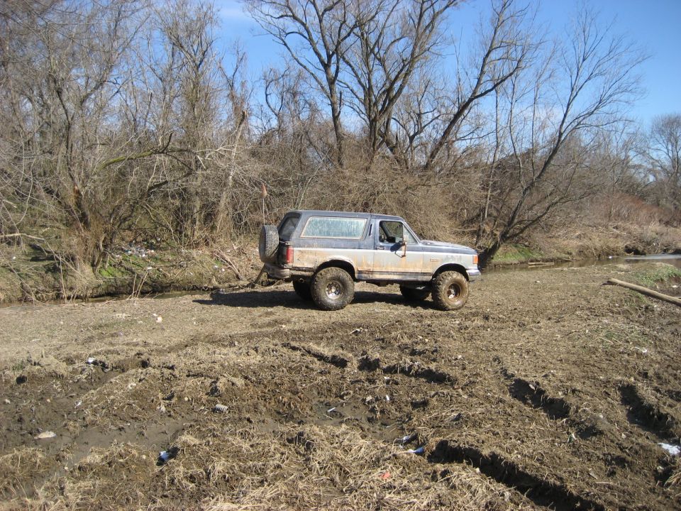Blue and silver Ford Bronco parked on muddy ground near a dry riverbed and bare trees.