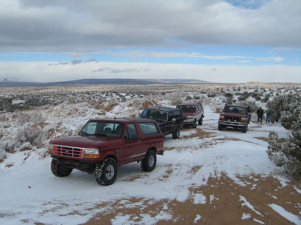 Red 1993 Ford Bronco on snowy dirt road with other vehicles and people.