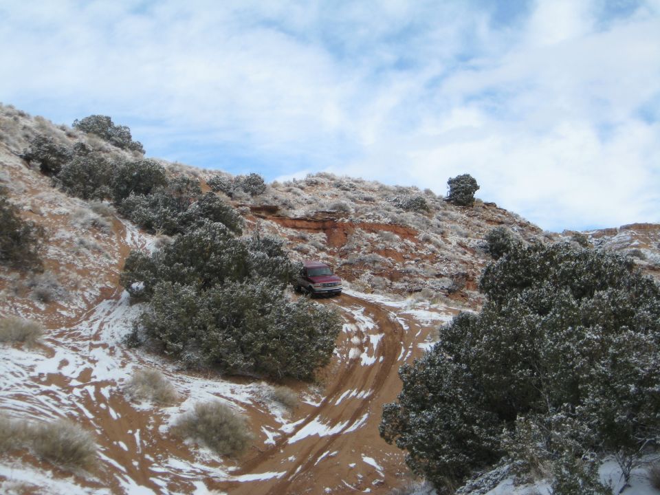 Red 1993 Ford Bronco driving on a snowy off-road trail up a sandy hill.