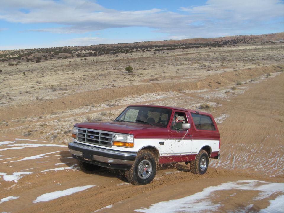 Red and white Ford Bronco driving on a snowy dirt road in a desert landscape.