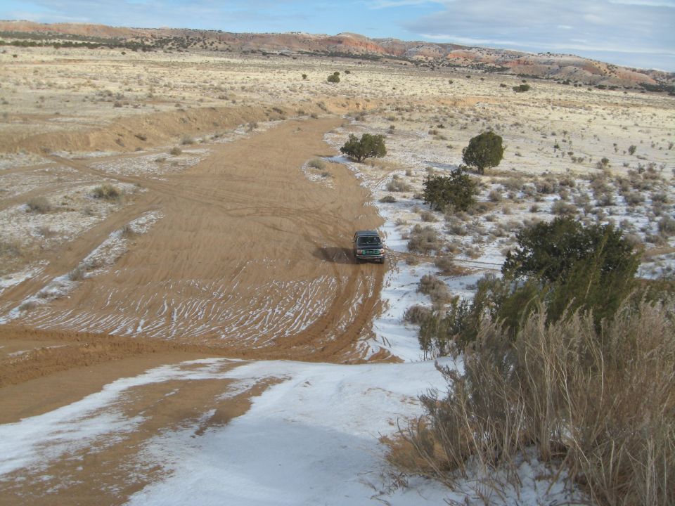 Dark SUV driving on a snowy dirt road in a desert landscape.