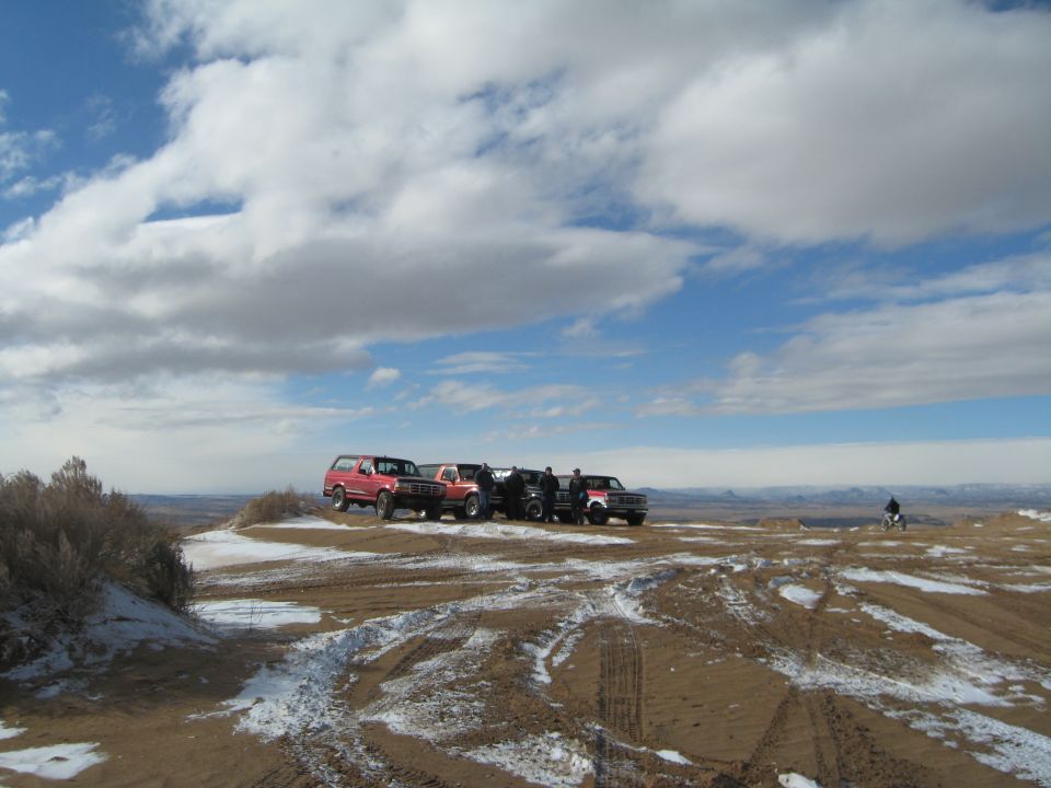 Red Ford Bronco and trucks on a hilltop with a motorcycle rider in the distance.